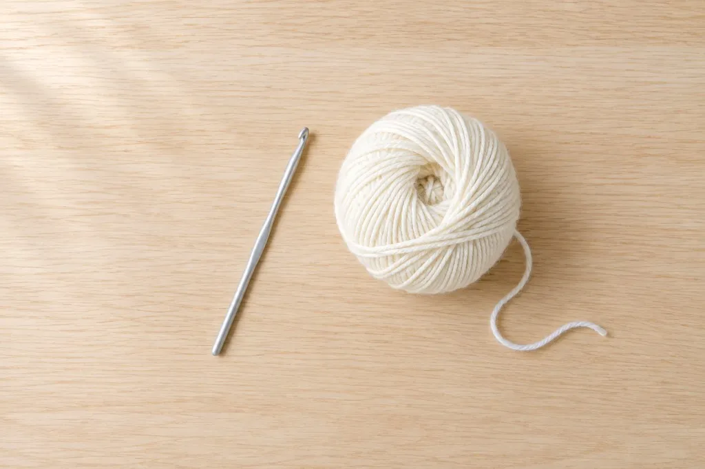 A single aluminum crochet hook beside one ball of ivory worsted-weight yarn